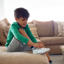 teaching self-sufficiency in children with special needs, avoiding learned helplessness, iStock image of Side view of Cute African american boy tying shoelace on sofa in living room at comfortable home