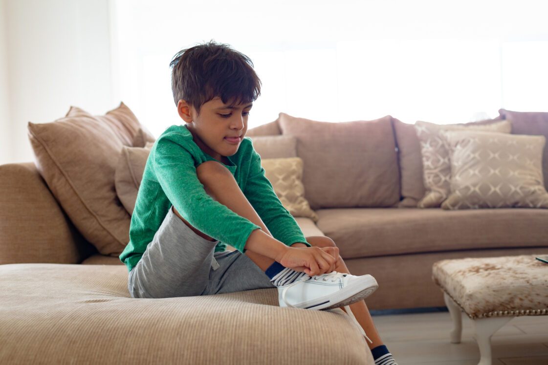 teaching self-sufficiency in children with special needs, avoiding learned helplessness, iStock image of Side view of Cute African american boy tying shoelace on sofa in living room at comfortable home
