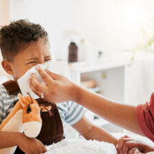 iStock image of: Flu, sick or cold child with parent sneezing, blowing and wiping runny nose while ill with covid virus, sinus and allergy symptoms in bed at home. Mother caring for stuffy and congested little son