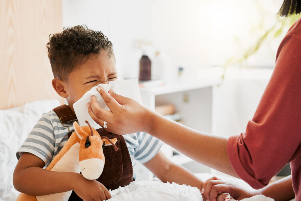 iStock image of: Flu, sick or cold child with parent sneezing, blowing and wiping runny nose while ill with covid virus, sinus and allergy symptoms in bed at home. Mother caring for stuffy and congested little son
