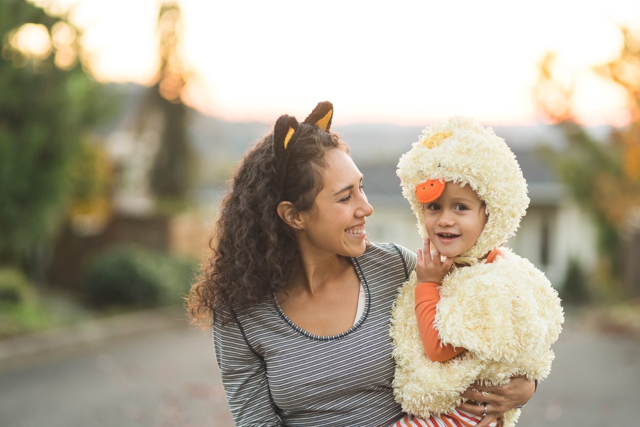 sensory friendly Halloween events, iStock images of a mom holds her toddler-age boy as he smiles at the camera while they're trick or treating. He's dressed in a duck costume, she has has cat ears on.