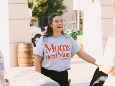 Image of a woman wearing a shirt that reads "Moms need moms" for an event with The Mom Walk Collective groups in Dallas-Fort Worth, photo courtesy @honeylandphoto